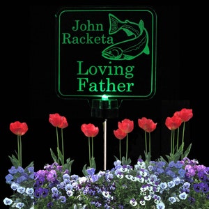 Pode incluir: Placa memorial verde iluminada com uma silhueta de peixe e o texto "John Racketa Loving Father". A placa est&aacute; montada em uma estaca de metal preto e cercada por violetas roxas e brancas e tulipas vermelhas.