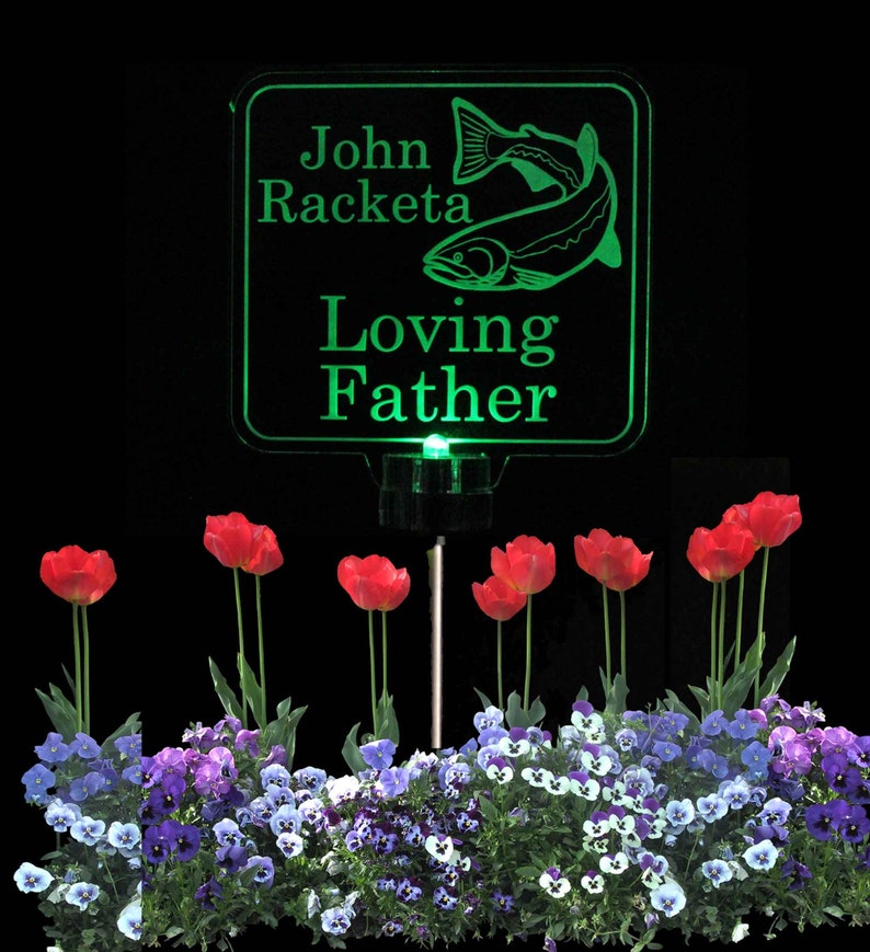 Pode incluir: Placa memorial verde iluminada com uma silhueta de peixe e o texto "John Racketa Loving Father". A placa est&aacute; montada em uma estaca de metal preto e cercada por violetas roxas e brancas e tulipas vermelhas.