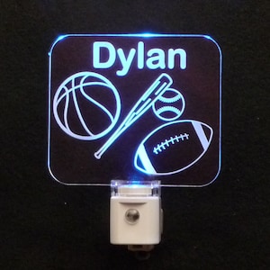 May include: A white acrylic night light with a blue LED light. The light features a basketball, baseball, bat, and football design. The name "Dylan" is written at the top of the light.