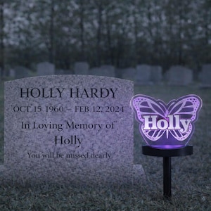 May include: A memorial scene featuring a headstone and a glowing butterfly-shaped solar light. The headstone reads "HOLLY HARDY" with dates and an inscription. The butterfly light displays the name "Holly".