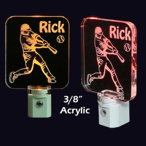 May include: Two 3/8 inch acrylic night lights with a baseball player silhouette and the name "Rick" engraved. The night light on the left is lit with a warm orange glow, while the night light on the right is lit with a pink glow.
