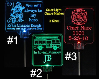 May include: Three solar-powered grave markers with different designs. The first marker is blue and features a firefighter with the text "You will always be my hero Ryan Charles Keogh February 14th 1979 - May 16th 2022". The second marker is green and features a fire truck with the text "In Loving Memory JB". The third marker is red and features a fire department logo with the text "Chief Nace 1101 5-23-10".