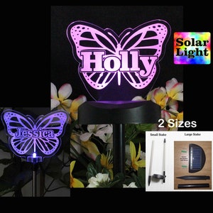 May include: A solar powered butterfly garden stake with the name "Holly" on it. The stake is made of metal and has a clear acrylic butterfly with a white outline. The butterfly is illuminated with a purple light. The stake is shown next to a smaller stake with the name "Jessica" on it. The stakes are available in two sizes, small and large. The image also shows the solar panel and stake for the large size.