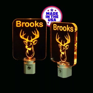 May include: Two white night lights with a deer silhouette and the name "Brooks" on each. The night lights are illuminated with a warm orange glow.