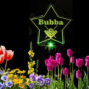 May include: A green illuminated star-shaped sign with the name "Bubba" and a pool cue with billiard balls. The sign is in front of a flower garden with red, pink, and yellow flowers.