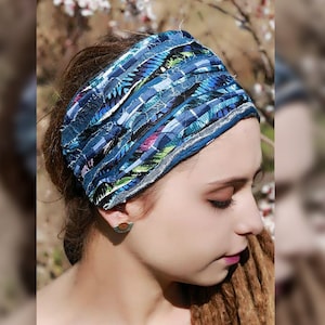 Op de afbeelding: Een brede stoffen hoofdband met een patchwork van blauwe, zwarte, witte en groen gemanipuleerde textielstroken. Het ontwerp bestaat uit meerdere horizontale lagen stof met gerafelde randen, wat zorgt voor een getextureerd uiterlijk over de band.