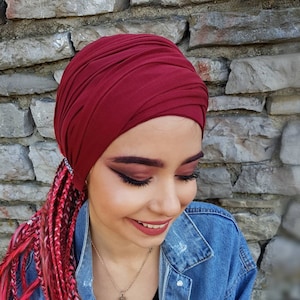 Può includere: Un turbante bordeaux. La persona indossa una giacca di jeans e ha lunghi capelli intrecciati rossi e rosa. È visibile una collana d'argento con un ciondolo blu.