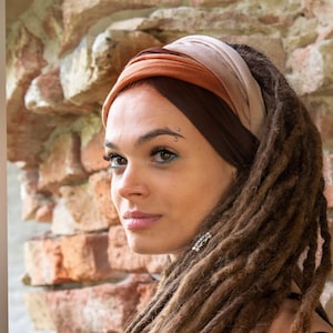 May include: A woman with long brown dreadlocks wears a brown and beige headband. The headband is made of soft fabric and has a simple design. The woman is looking to the side and has a neutral expression.