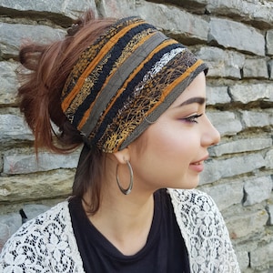 Op de afbeelding: Een brede stoffen hoofdband met een patchworkontwerp van zwarte, mosterdgele en grijze horizontale strepen met ingewikkelde kanten en gestructureerde stoffen overlays. Het accessoire is aan de achterkant gestrikt en toont een mix van effen en patronen.