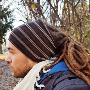 May include: A brown beanie with thin horizontal white stripes worn over long brown dreadlocks. The headwear is paired with a white fringed scarf and a black jacket with a blue inner layer, featuring a small black nose ring on the wearer.