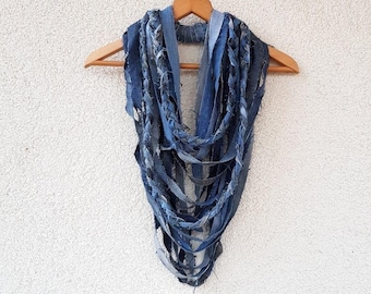May include: A scarf constructed from multiple strips of blue denim fabric in varying shades of indigo and light blue. The strips are arranged in a layered, looped design with frayed, raw edges, creating a textured, multi-strand appearance.