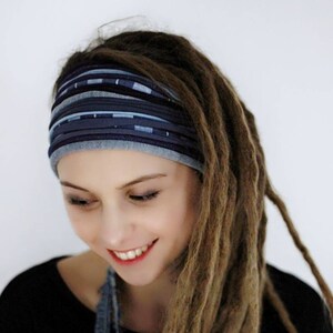 Op de afbeelding: Een brede stoffen haarband met horizontale strepen in diverse blauwtinten, waaronder marineblauw, spijkerblauw en lichtblauw, met een textuur. De haarband wordt over het voorhoofd gedragen en vormt een contrast met lange, lichtbruine dreadlocks.