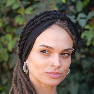 Könnte beinhalten: Ein schwarzer Turban-Stil-Kopftuch mit einem geflochtenen Design. Das Kopftuch ist aus einem weichen Stoff gefertigt und wird auf dem Kopf getragen. Der Hintergrund ist ein verschwommenes grünes Laub.