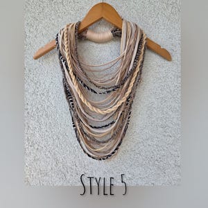 Op de afbeelding: Een meerlagige ketting in beige, lichtbeige en zwart, hangend aan een houten hanger. De ketting heeft gevlochten en gefranjerde elementen. De tekst "STYLE 5" is onderaan zichtbaar.