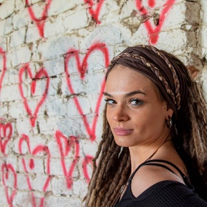 May include: A dark brown fabric headband featuring a braided design with metallic accents. The headband is worn across the forehead, securing long dreadlocked hair. The accessory is constructed from a smooth, flexible material with a wide band.
