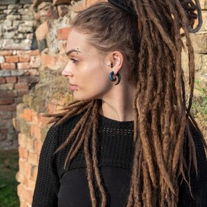 May include: A dark blue spiral ear gauge earring worn in a pierced earlobe. The jewelry features a curved, tapered design with a smooth finish, showcasing a deep blue hue with subtle dark swirls, positioned against a profile view of a person.
