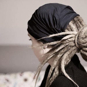 K&ouml;nnte beinhalten: Eine Person mit Dreadlocks tr&auml;gt ein dunkelblaues Kopftuch. Die Dreadlocks sind zu einem Dutt gestylt. Die Person tr&auml;gt ein schwarzes Oberteil. Der Hintergrund ist neutral gef&auml;rbt.