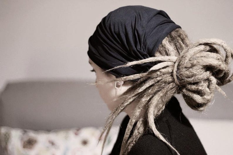 K&ouml;nnte beinhalten: Eine Person mit Dreadlocks tr&auml;gt ein dunkelblaues Kopftuch. Die Dreadlocks sind zu einem Dutt gestylt. Die Person tr&auml;gt ein schwarzes Oberteil. Der Hintergrund ist neutral gef&auml;rbt.