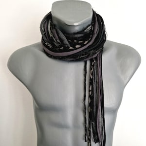 May include: A multi-strand scarf featuring a combination of black, charcoal gray, and light gray fabric strips. The scarf is constructed from various textures, including solid colors and a subtle spotted pattern, with frayed edges hanging down.