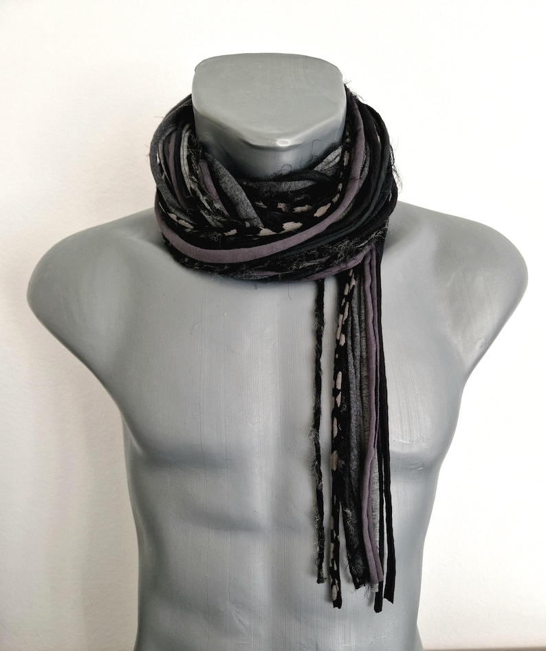 May include: A multi-strand scarf featuring a combination of black, charcoal gray, and light gray fabric strips. The scarf is constructed from various textures, including solid colors and a subtle spotted pattern, with frayed edges hanging down.