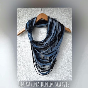 Può includere: Una sciarpa in denim fatta a mano, nei toni del blu e del nero, appesa a una gruccia di legno. La sciarpa è realizzata con strisce di tessuto denim, creando un design aperto e sfrangiato. Il testo "UNIKATINA DENIM SCARVES" è in basso.