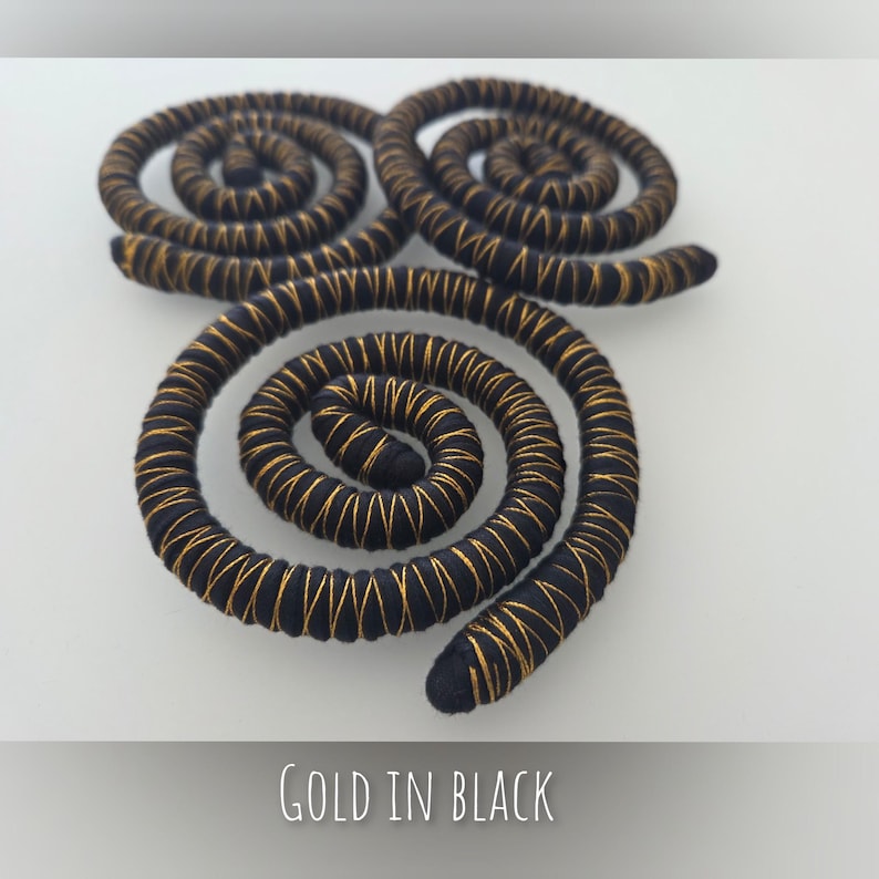 K&ouml;nnte beinhalten: Drei spiralf&ouml;rmige Dekorationselemente aus schwarzer Kordel, die mit d&uuml;nnem goldenem Metallic-Faden umwickelt sind. Die Spiralen sind in einer Dreiecksform angeordnet. Der Text "GOLD IN BLACK" steht in Wei&szlig; am unteren Bildrand.