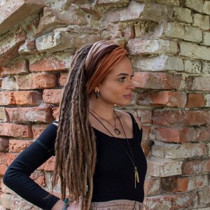 K&ouml;nnte beinhalten: Eine Frau mit langen braunen Dreadlocks tr&auml;gt ein braunes und orangefarbenes Samtband. Sie tr&auml;gt ein schwarzes Oberteil und einen braunen Rock. Sie steht vor einer Backsteinmauer.