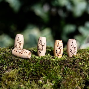 May include: Five wooden beads with the Om symbol carved into them. The beads are arranged in a row on a bed of green moss.