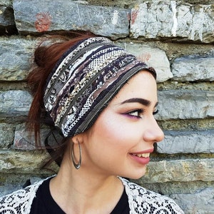 May include: A multi-colored headband with a patchwork design. The headband features various patterns and textures in shades of brown, gray, and white. The accessory is worn on the head and is set against a stone wall background.