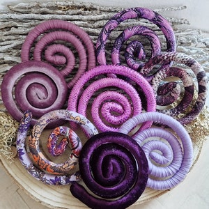 A collection of eight purple and pink fabric coiled spirals. The spirals are made of different fabrics, including velvet, satin, and patterned fabric. The spirals are arranged in a circle on a wooden surface.