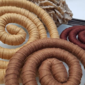 May include: Three spiral-shaped decorative objects wrapped in fabric in shades of light tan, medium brown, and dark chocolate brown. Each spiral features a consistent ribbed texture created by the fabric wrapping, arranged in a circular coil pattern.