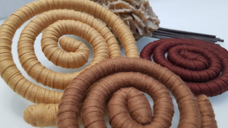 May include: Three spiral-shaped decorative objects wrapped in fabric in shades of light tan, medium brown, and dark chocolate brown. Each spiral features a consistent ribbed texture created by the fabric wrapping, arranged in a circular coil pattern.