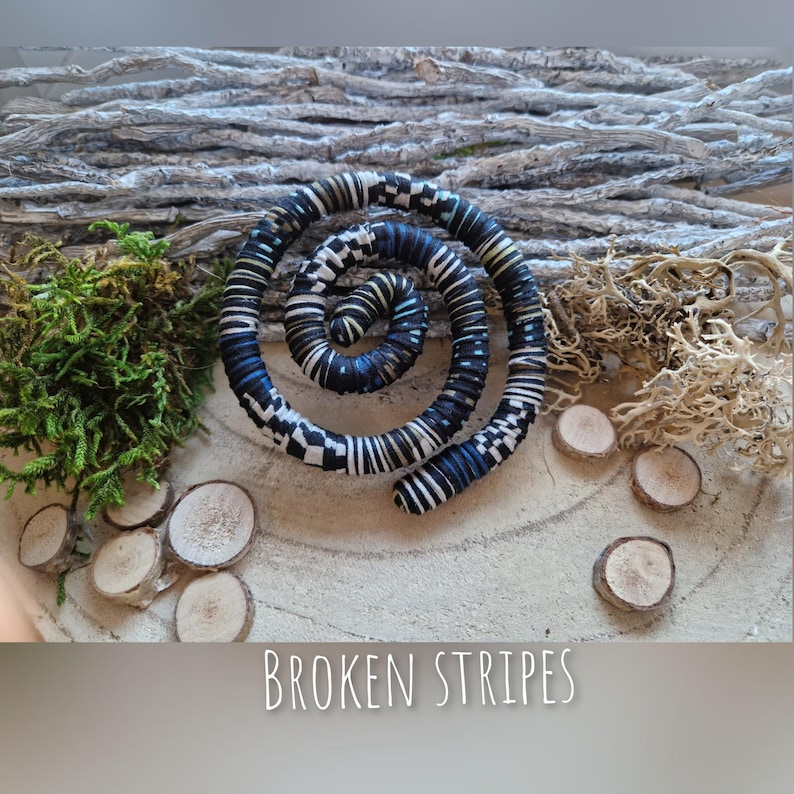 May include: A spiral-shaped decorative item with a black, white, and blue broken stripe pattern. The item is centered on a wooden surface, surrounded by moss, wood slices, and twigs. The text "BROKEN STRIPES" is at the bottom.
