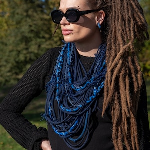 Puede incluir: Bufanda azul oscuro hecha a mano con un diseño en capas. La bufanda está hecha de tiras de tela en varios tonos de azul. La persona de la imagen lleva gafas de sol negras, una camiseta negra y tiene largas rastas.