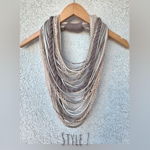 Op de afbeelding: Een multi-strand infinity sjaal in tinten grijs, taupe en cr&egrave;me, weergegeven op een houten hanger. De sjaal heeft verschillende texturen, waaronder gevlochten en geweven elementen. De tekst "STYLE 7" is zichtbaar onderaan.
