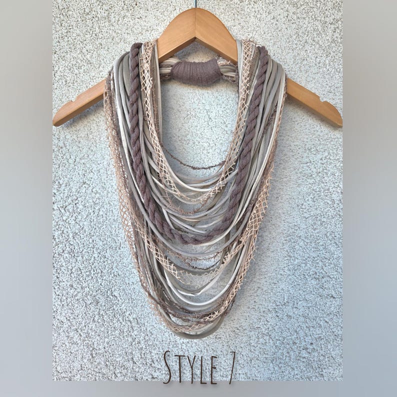 Op de afbeelding: Een multi-strand infinity sjaal in tinten grijs, taupe en cr&egrave;me, weergegeven op een houten hanger. De sjaal heeft verschillende texturen, waaronder gevlochten en geweven elementen. De tekst "STYLE 7" is zichtbaar onderaan.