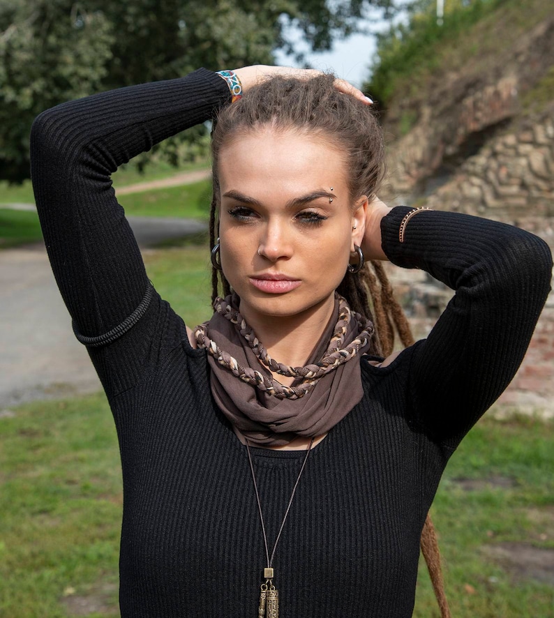 May include: A brown infinity scarf featuring a braided texture with lighter brown and cream accents. The scarf is worn around the neck, showcasing a soft fabric construction with multiple intertwined strands that create a layered, circular appearance.