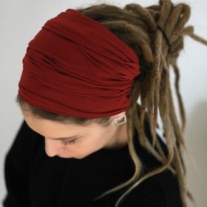 K&ouml;nnte beinhalten: Ein tiefrotes Stirnband, wahrscheinlich aus Stoff, bedeckt den oberen Teil des Kopfes. Das Stirnband ist mit mehreren Falten versehen. Lange, braune Dreadlocks erstrecken sich unter dem Stirnband. Die Person tr&auml;gt ein schwarzes Oberteil.
