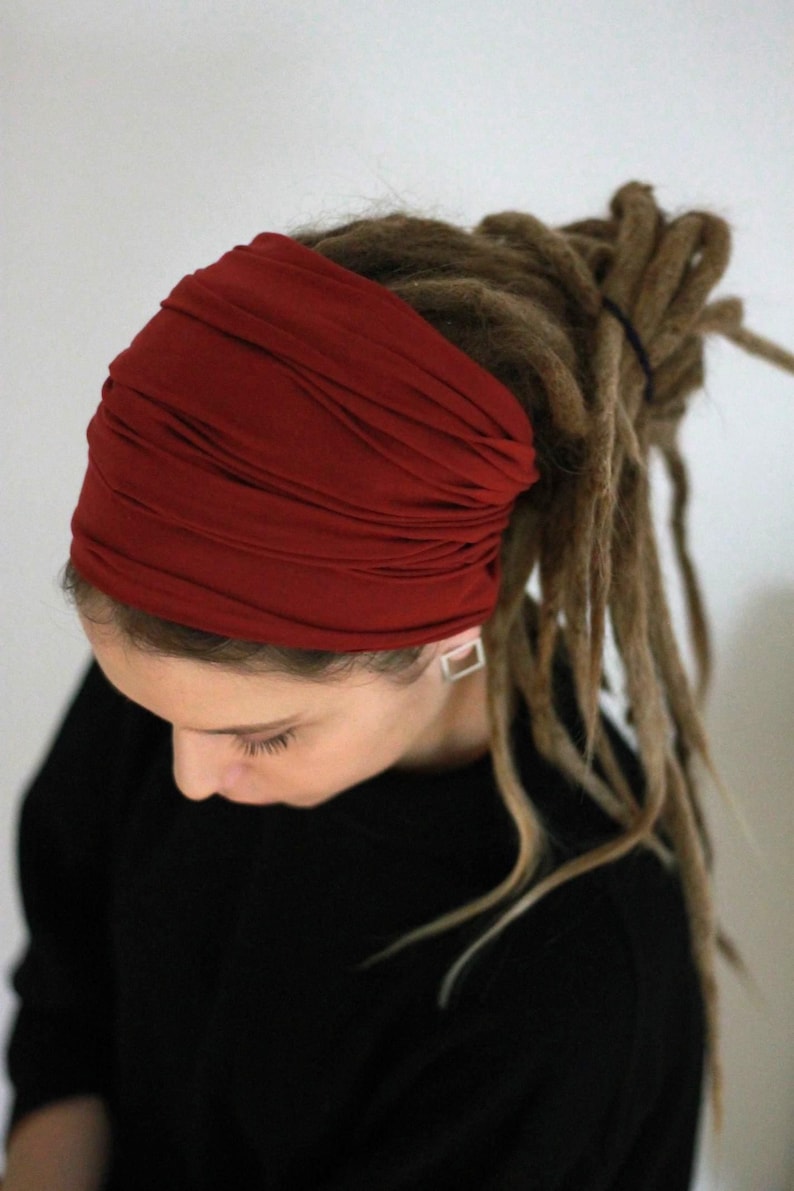 K&ouml;nnte beinhalten: Ein tiefrotes Stirnband, wahrscheinlich aus Stoff, bedeckt den oberen Teil des Kopfes. Das Stirnband ist mit mehreren Falten versehen. Lange, braune Dreadlocks erstrecken sich unter dem Stirnband. Die Person tr&auml;gt ein schwarzes Oberteil.