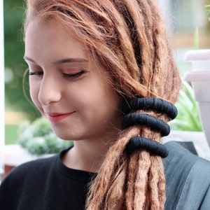 May include: Three black fabric-covered hair ties wrapped around a section of long, light brown dreadlocks. The hair ties are cylindrical, textured, and positioned horizontally in a stack to secure the hair in a ponytail style.