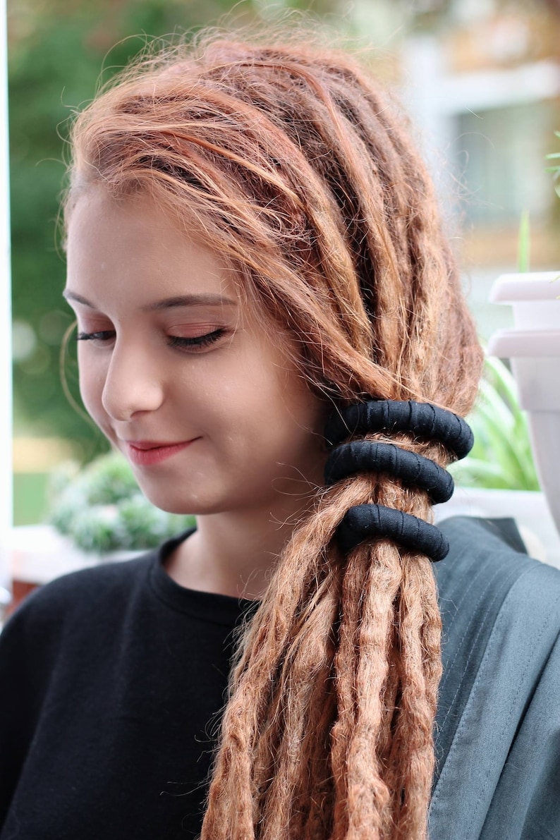 May include: Three black fabric-covered hair ties wrapped around a section of long, light brown dreadlocks. The hair ties are cylindrical, textured, and positioned horizontally in a stack to secure the hair in a ponytail style.