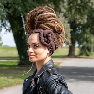 May include: A woman with brown dreadlocks and a dark brown head wrap. She is wearing a black leather jacket and hoop earrings. The background shows a park with trees and a paved path.