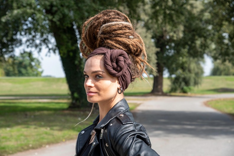 May include: A woman with brown dreadlocks and a dark brown head wrap. She is wearing a black leather jacket and hoop earrings. The background shows a park with trees and a paved path.