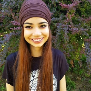 May include: A dark brown turban-style head wrap. The person is wearing a black t-shirt with a white graphic design. The background features green and purple foliage. The person has long auburn hair and is smiling.