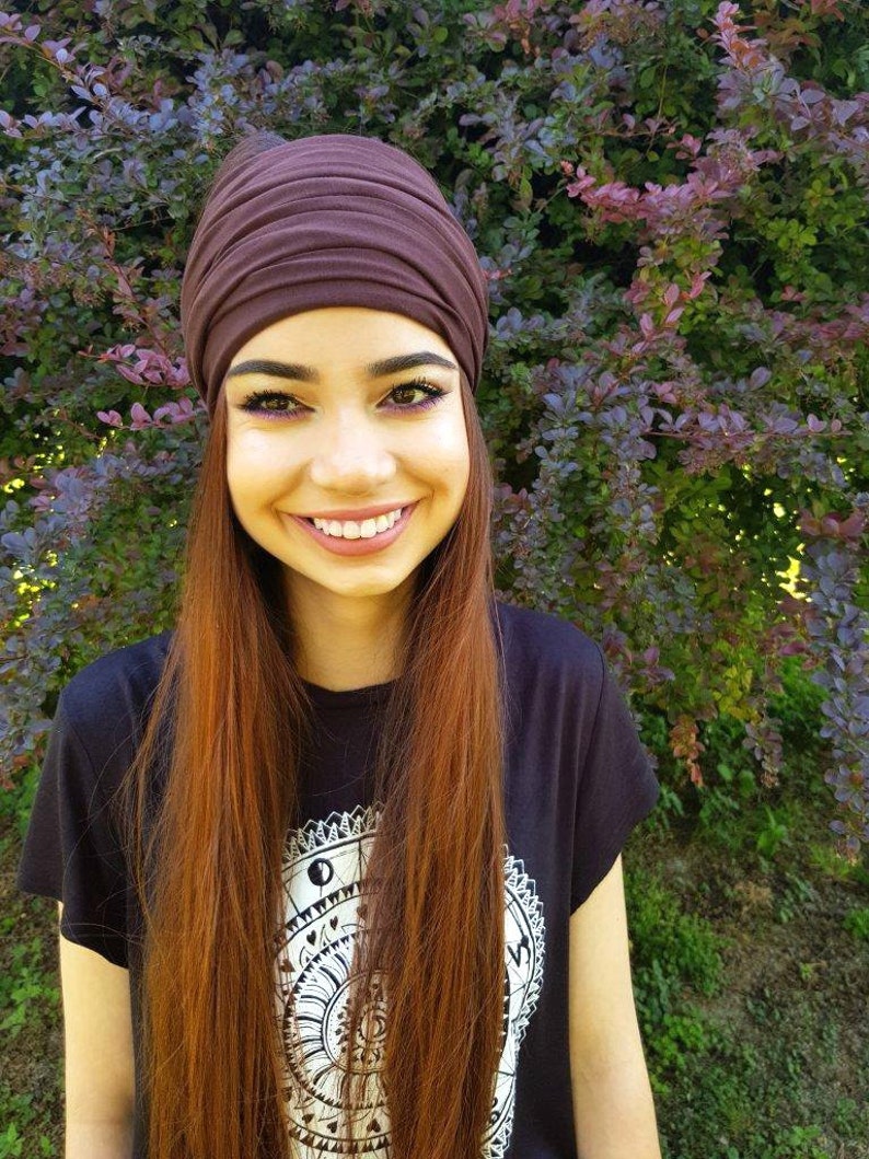 May include: A dark brown turban-style head wrap. The person is wearing a black t-shirt with a white graphic design. The background features green and purple foliage. The person has long auburn hair and is smiling.