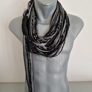 May include: A multi-strand infinity scarf featuring a mix of black, charcoal gray, and light gray fabric strips. The scarf includes textured, frayed edges and a solid black fabric-wrapped accent section, creating a layered, voluminous appearance.