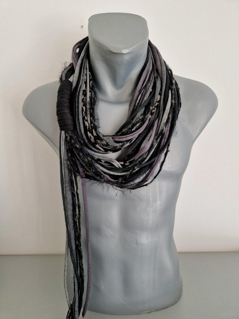 May include: A multi-strand infinity scarf featuring a mix of black, charcoal gray, and light gray fabric strips. The scarf includes textured, frayed edges and a solid black fabric-wrapped accent section, creating a layered, voluminous appearance.