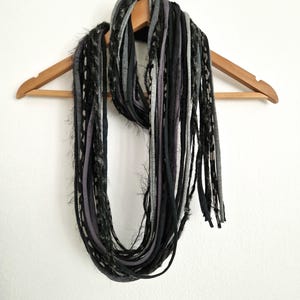 May include: A multi-strand infinity scarf composed of numerous long, thin fabric strips in varying shades of black, charcoal gray, and light gray. The scarf features a mix of smooth jersey-like textures and frayed, fuzzy yarn-like strands.