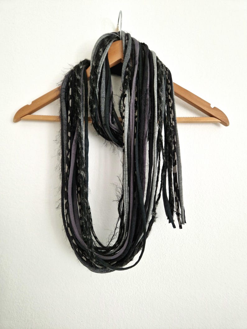 May include: A multi-strand infinity scarf composed of numerous long, thin fabric strips in varying shades of black, charcoal gray, and light gray. The scarf features a mix of smooth jersey-like textures and frayed, fuzzy yarn-like strands.