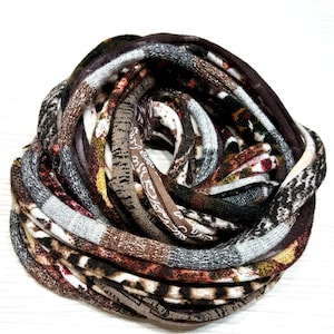 May include: A circular infinity scarf featuring a multi-patterned knit fabric in shades of brown, black, white, and gray. The textile displays various textures and prints, including animal-inspired motifs and geometric patterns, coiled into a dense, layered loop.